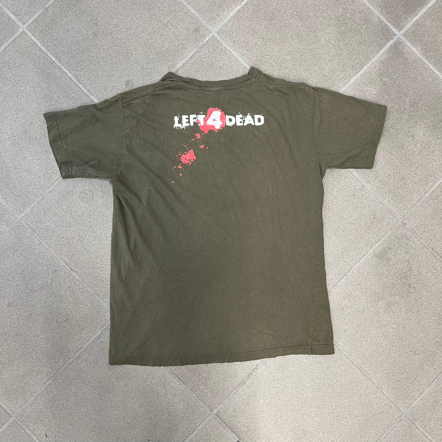 Left 4 Dead 2009 Infection Warning Tee features a khaki T-shirt with a distressed poster-style graphic on the front. The graphic has red “Infection Warning” text, instructions on what to do when infected, an emergency hotline number, and scribbled details. A Left 4 Dead logo and small 2009 Valve licensing appear underneath. The upper back features a Left 4 Dead logo with a blood splatter graphic.