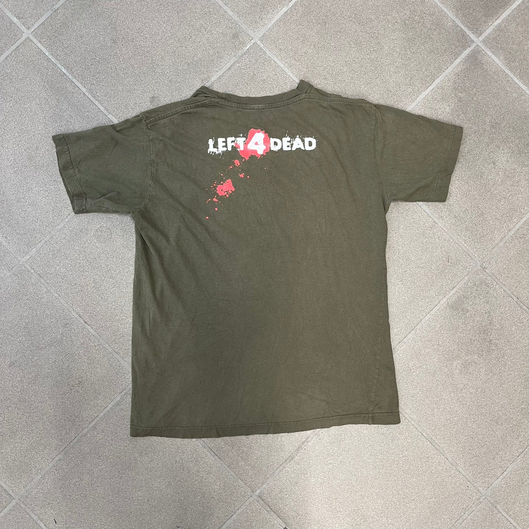 Left 4 Dead 2009 Infection Warning Tee features a khaki T-shirt with a distressed poster-style graphic on the front. The graphic has red “Infection Warning” text, instructions on what to do when infected, an emergency hotline number, and scribbled details. A Left 4 Dead logo and small 2009 Valve licensing appear underneath. The upper back features a Left 4 Dead logo with a blood splatter graphic.