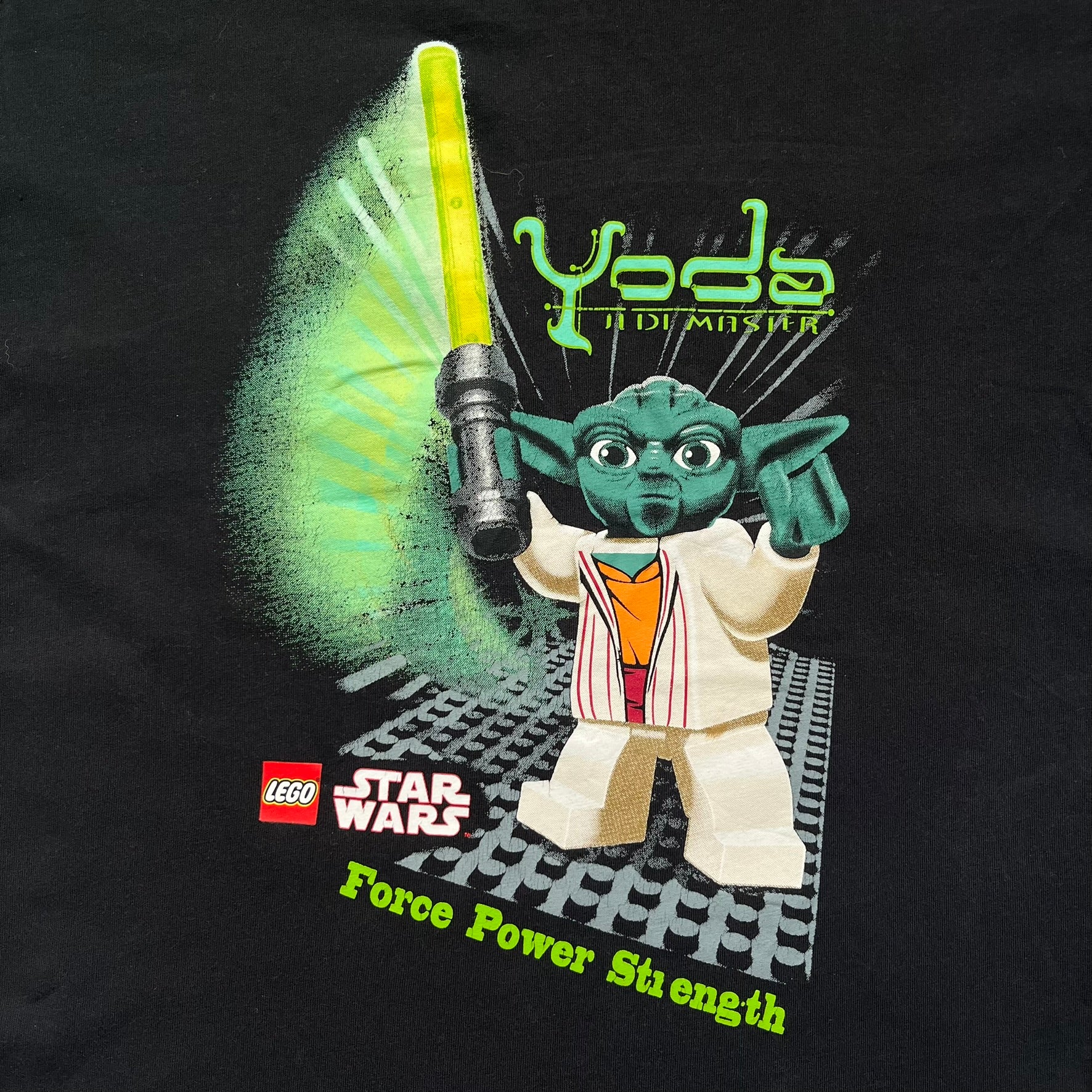 LEGO Yoda “Jedi Master” 2009 Tee is a black short-sleeve t-shirt featuring a front graphic of Yoda swinging a green lightsaber. Above the graphic is “Yoda Jedi Master” text in green, with “Force Power Strength” printed underneath. The tee includes a LEGO Star Wars 2009 neck tag.