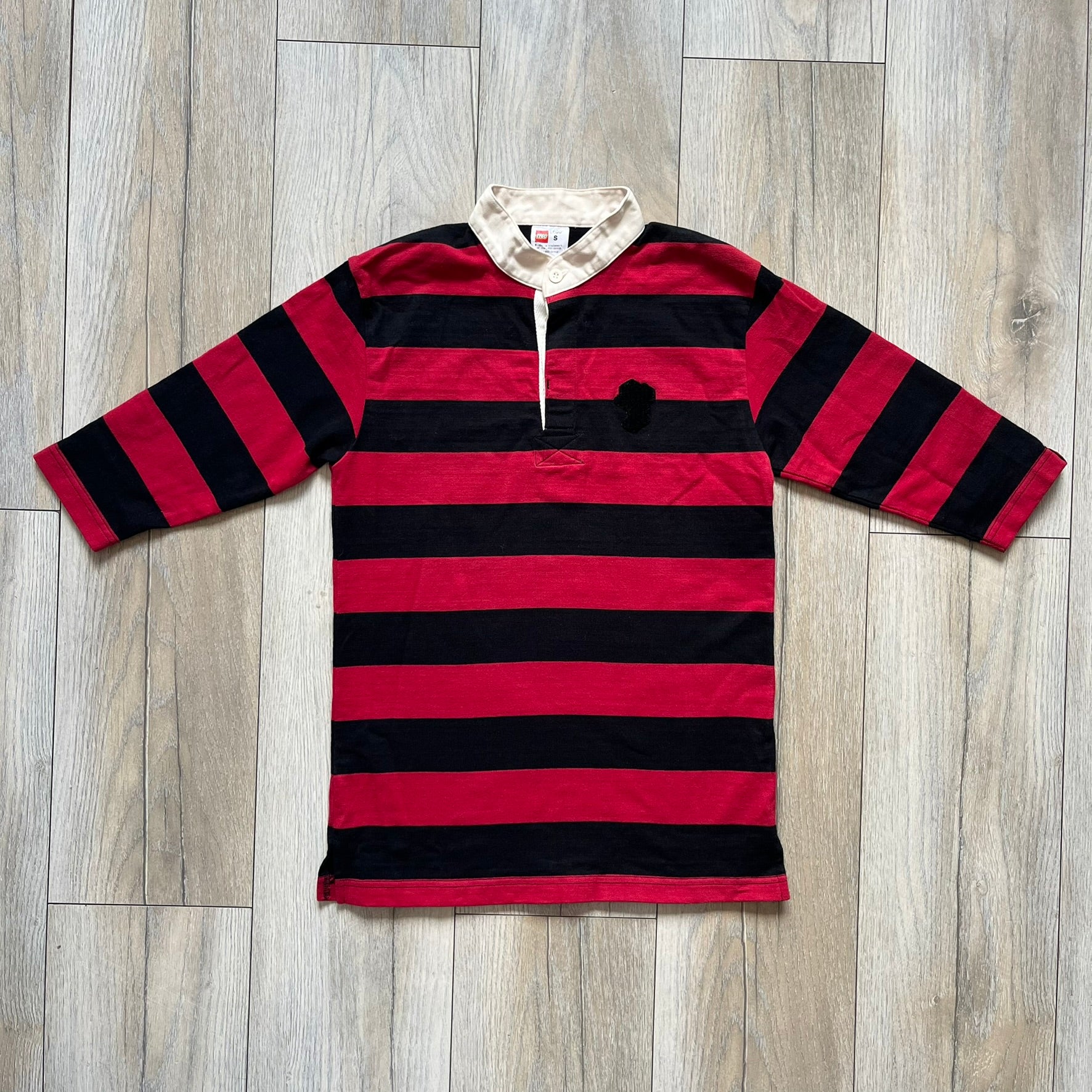 LEGO 2000 Black &amp; Red Rugby Shirt is a black and red striped long-sleeve rugby shirt with a cream/white collar. The chest includes a black Velcro patch panel without an attached patch, and a small LEGO tag is placed on one of the sleeves. The shirt has an official LEGO 2000 licensed neck tag.