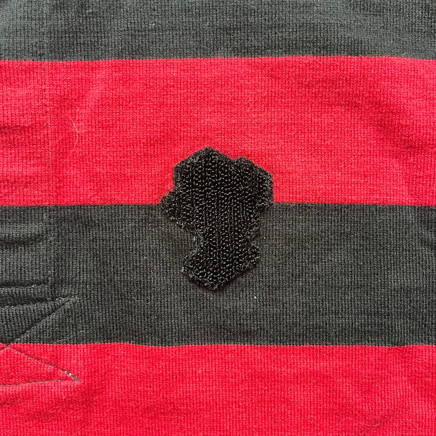 LEGO 2000 Black &amp; Red Rugby Shirt is a black and red striped long-sleeve rugby shirt with a cream/white collar. The chest includes a black Velcro patch panel without an attached patch, and a small LEGO tag is placed on one of the sleeves. The shirt has an official LEGO 2000 licensed neck tag.