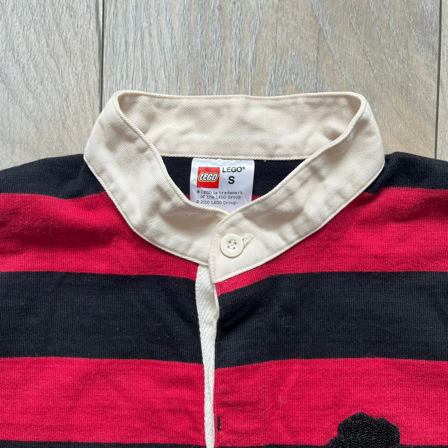 LEGO 2000 Black &amp; Red Rugby Shirt is a black and red striped long-sleeve rugby shirt with a cream/white collar. The chest includes a black Velcro patch panel without an attached patch, and a small LEGO tag is placed on one of the sleeves. The shirt has an official LEGO 2000 licensed neck tag.