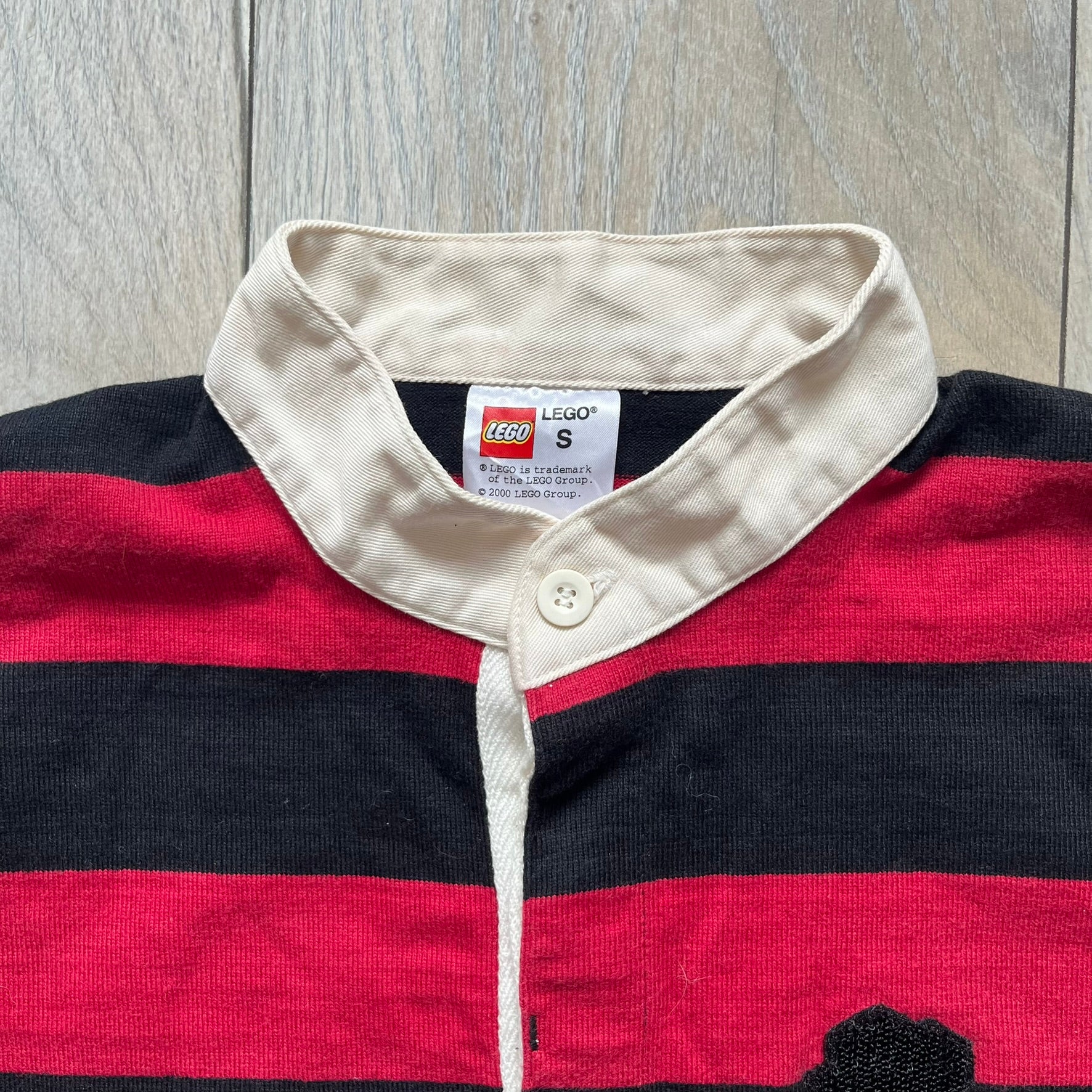 LEGO 2000 Black &amp; Red Rugby Shirt is a black and red striped long-sleeve rugby shirt with a cream/white collar. The chest includes a black Velcro patch panel without an attached patch, and a small LEGO tag is placed on one of the sleeves. The shirt has an official LEGO 2000 licensed neck tag.