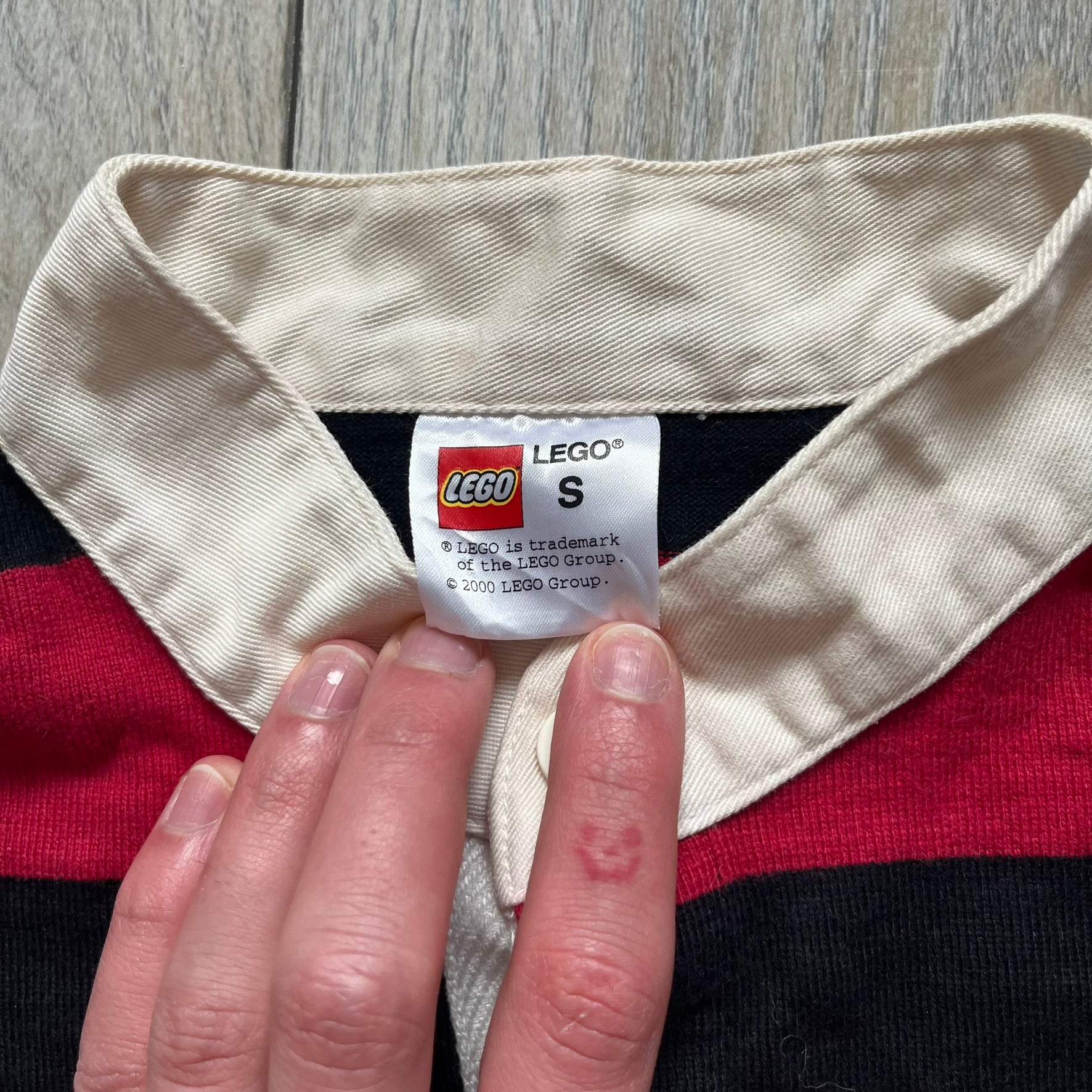 LEGO 2000 Black &amp; Red Rugby Shirt is a black and red striped long-sleeve rugby shirt with a cream/white collar. The chest includes a black Velcro patch panel without an attached patch, and a small LEGO tag is placed on one of the sleeves. The shirt has an official LEGO 2000 licensed neck tag.