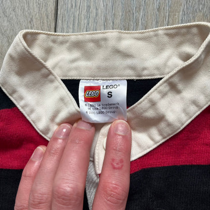 LEGO 2000 Black &amp; Red Rugby Shirt is a black and red striped long-sleeve rugby shirt with a cream/white collar. The chest includes a black Velcro patch panel without an attached patch, and a small LEGO tag is placed on one of the sleeves. The shirt has an official LEGO 2000 licensed neck tag.