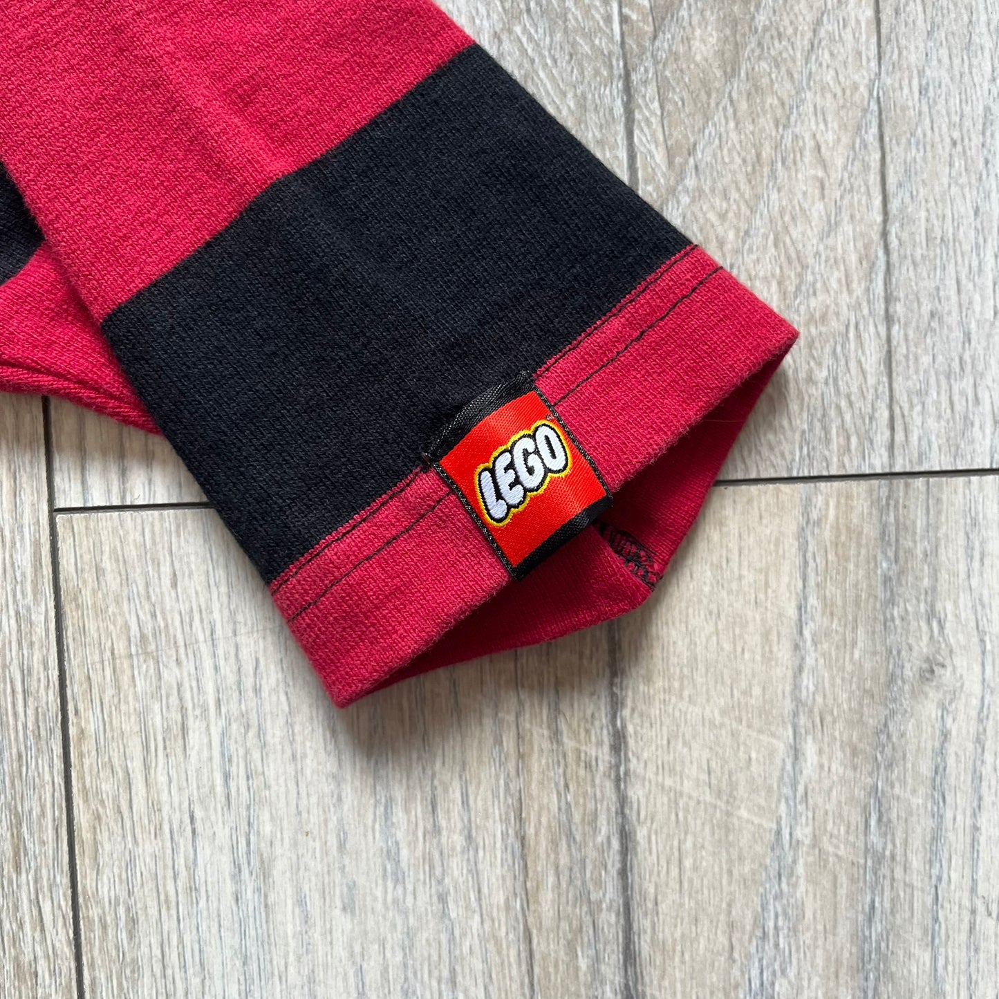 LEGO 2000 Black &amp; Red Rugby Shirt is a black and red striped long-sleeve rugby shirt with a cream/white collar. The chest includes a black Velcro patch panel without an attached patch, and a small LEGO tag is placed on one of the sleeves. The shirt has an official LEGO 2000 licensed neck tag.