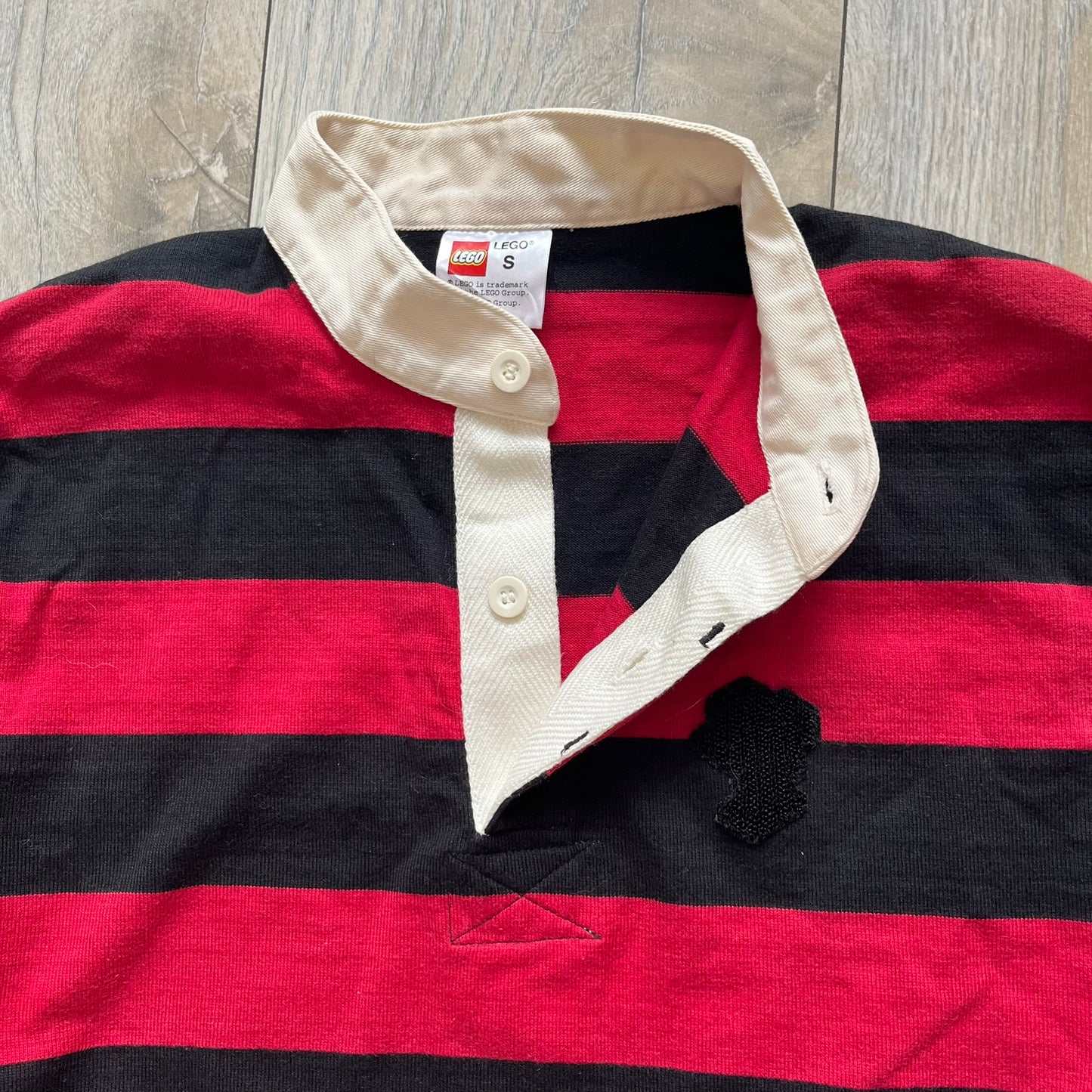 LEGO 2000 Black &amp; Red Rugby Shirt is a black and red striped long-sleeve rugby shirt with a cream/white collar. The chest includes a black Velcro patch panel without an attached patch, and a small LEGO tag is placed on one of the sleeves. The shirt has an official LEGO 2000 licensed neck tag.
