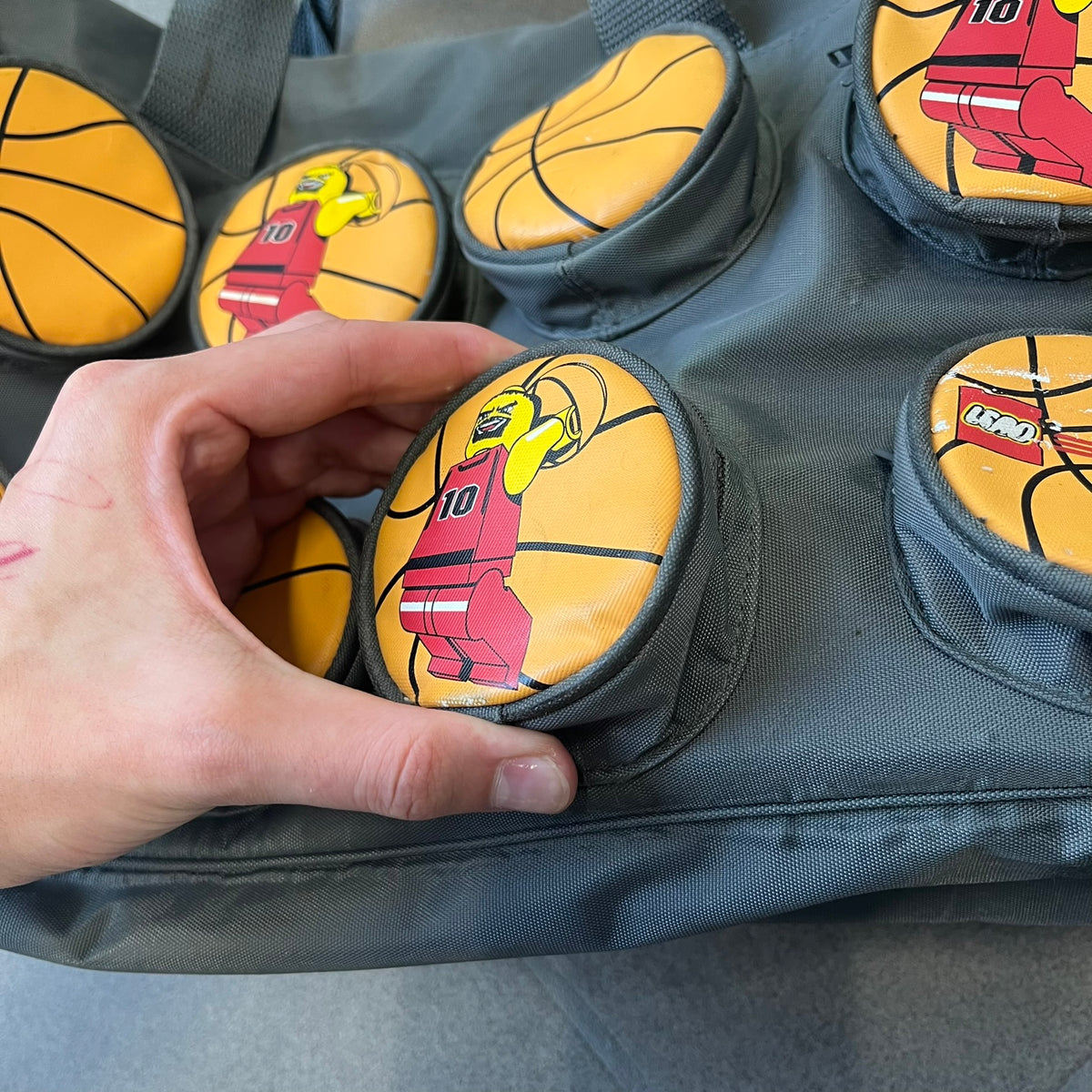 Lego Basketball 2003 Multi Pocket Gray Bag is a gray gym bag with orange side panels and an orange strap handle, featuring a Lego Sports tag. The front is designed to resemble a Lego 8x2 brick, with 8 zippable circular pockets featuring basketball orange graphics or basketball player designs.