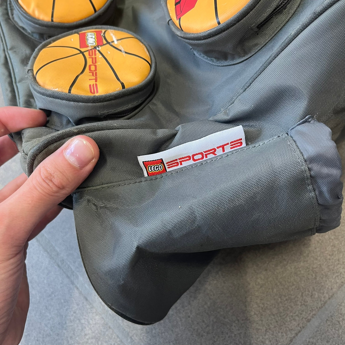 Lego Basketball 2003 Multi Pocket Gray Bag is a gray gym bag with orange side panels and an orange strap handle, featuring a Lego Sports tag. The front is designed to resemble a Lego 8x2 brick, with 8 zippable circular pockets featuring basketball orange graphics or basketball player designs.