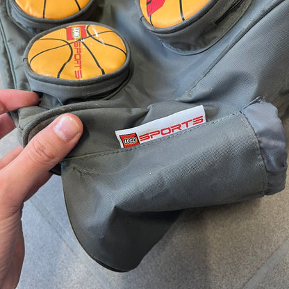 Lego Basketball 2003 Multi Pocket Gray Bag is a gray gym bag with orange side panels and an orange strap handle, featuring a Lego Sports tag. The front is designed to resemble a Lego 8x2 brick, with 8 zippable circular pockets featuring basketball orange graphics or basketball player designs.