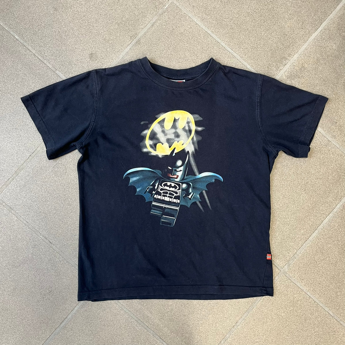 Lego Batman 2006 TS39 Promo Tee is a black T-shirt released alongside the first Lego Batman sets in 2006, featuring a Lego Batman graphic on the front and a clouded Bat-Signal on the back. A licensed 2006 Lego Batman patch sits on the upper back, and it has a Lego Collection neck tag and a small Lego tag on the side. This tee has no size tag.