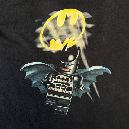 Lego Batman 2006 TS39 Promo Tee is a black T-shirt released alongside the first Lego Batman sets in 2006, featuring a Lego Batman graphic on the front and a clouded Bat-Signal on the back. A licensed 2006 Lego Batman patch sits on the upper back, and it has a Lego Collection neck tag and a small Lego tag on the side. This tee has no size tag.