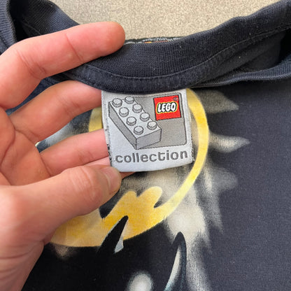 Lego Batman 2006 TS39 Promo Tee is a black T-shirt released alongside the first Lego Batman sets in 2006, featuring a Lego Batman graphic on the front and a clouded Bat-Signal on the back. A licensed 2006 Lego Batman patch sits on the upper back, and it has a Lego Collection neck tag and a small Lego tag on the side. This tee has no size tag.