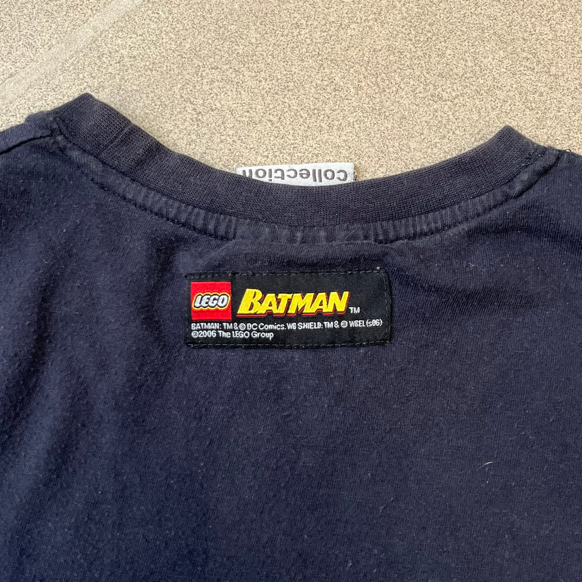 Lego Batman 2006 TS39 Promo Tee is a black T-shirt released alongside the first Lego Batman sets in 2006, featuring a Lego Batman graphic on the front and a clouded Bat-Signal on the back. A licensed 2006 Lego Batman patch sits on the upper back, and it has a Lego Collection neck tag and a small Lego tag on the side. This tee has no size tag.