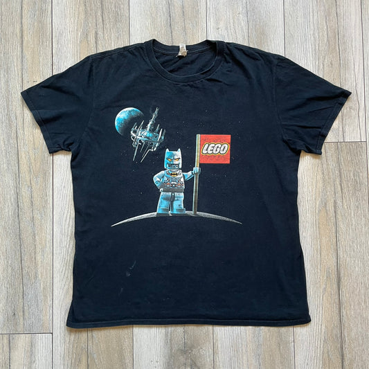 Lego Batman 3: Beyond Gotham 2014 Tee is a black T-shirt made on an Anvil blank. The front features a graphic of Batman in a white and blue suit standing on the moon, holding a Lego flag, with a planet and space station in the background. The back includes the “Batman 3: Beyond Gotham” logo and a Lego logo placed on the upper back area, with small licensing print beneath.