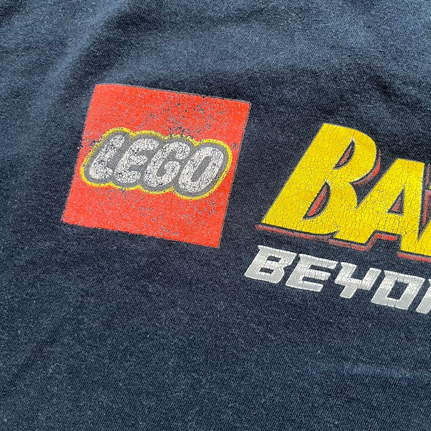 Lego Batman 3: Beyond Gotham 2014 Tee is a black T-shirt made on an Anvil blank. The front features a graphic of Batman in a white and blue suit standing on the moon, holding a Lego flag, with a planet and space station in the background. The back includes the “Batman 3: Beyond Gotham” logo and a Lego logo placed on the upper back area, with small licensing print beneath.