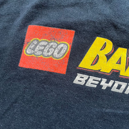 Lego Batman 3: Beyond Gotham 2014 Tee is a black T-shirt made on an Anvil blank. The front features a graphic of Batman in a white and blue suit standing on the moon, holding a Lego flag, with a planet and space station in the background. The back includes the “Batman 3: Beyond Gotham” logo and a Lego logo placed on the upper back area, with small licensing print beneath.