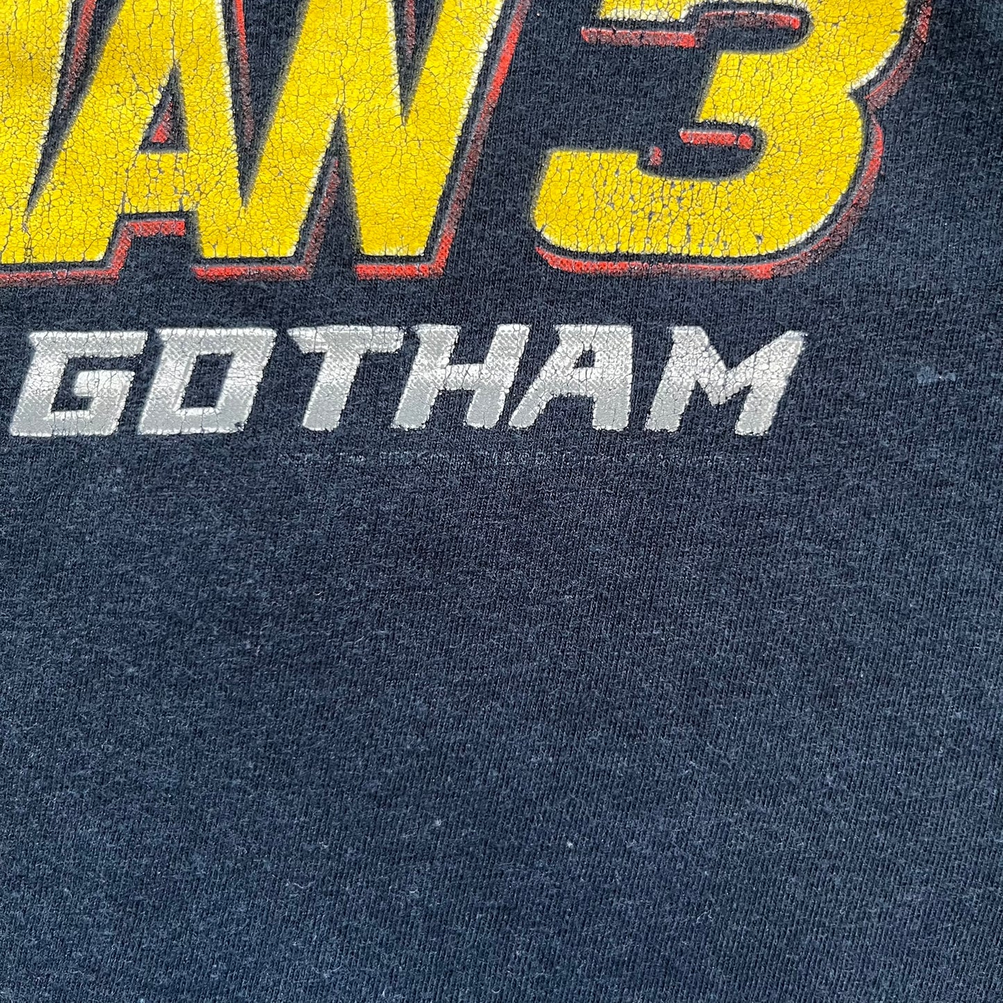 Lego Batman 3: Beyond Gotham 2014 Tee is a black T-shirt made on an Anvil blank. The front features a graphic of Batman in a white and blue suit standing on the moon, holding a Lego flag, with a planet and space station in the background. The back includes the “Batman 3: Beyond Gotham” logo and a Lego logo placed on the upper back area, with small licensing print beneath.