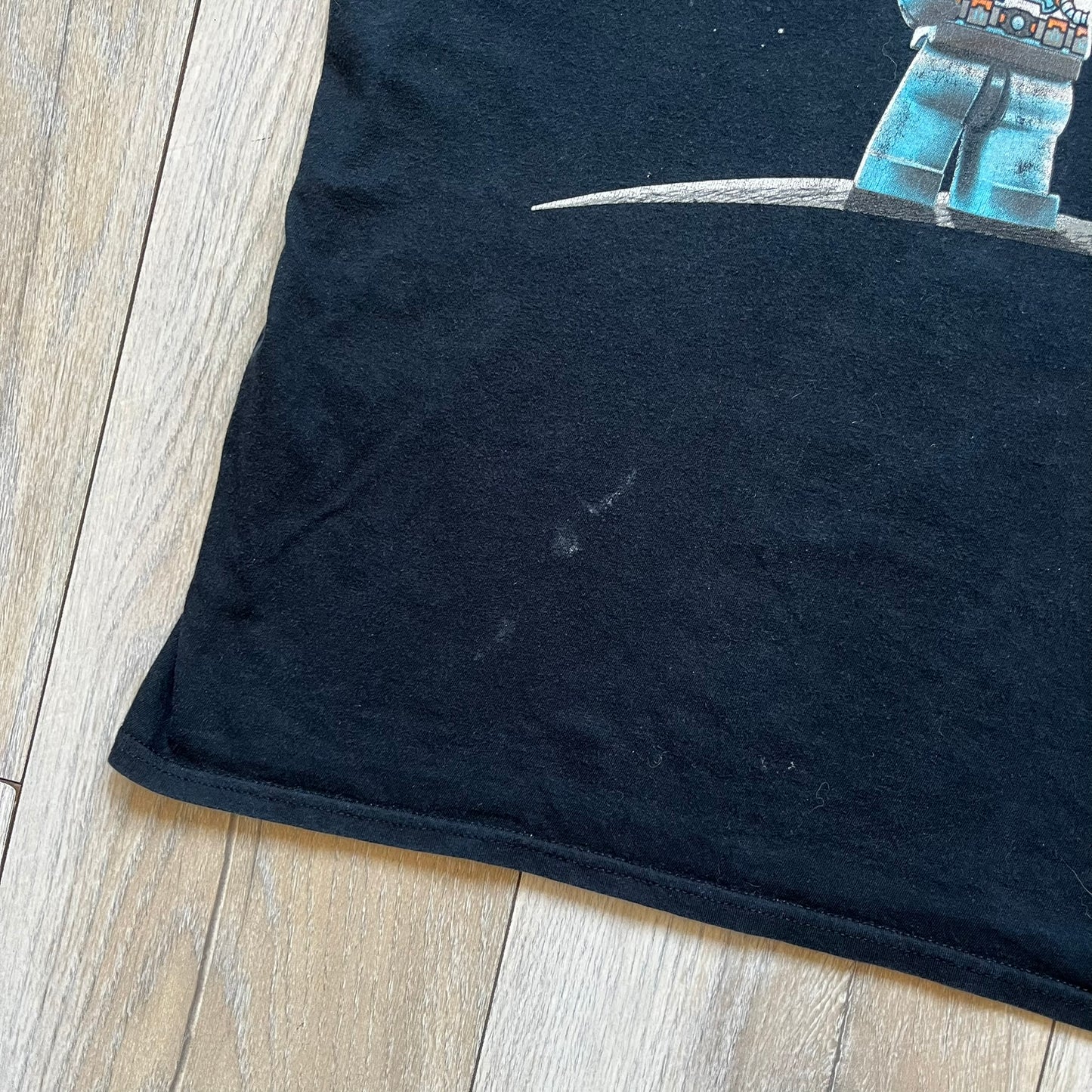 Lego Batman 3: Beyond Gotham 2014 Tee is a black T-shirt made on an Anvil blank. The front features a graphic of Batman in a white and blue suit standing on the moon, holding a Lego flag, with a planet and space station in the background. The back includes the “Batman 3: Beyond Gotham” logo and a Lego logo placed on the upper back area, with small licensing print beneath.