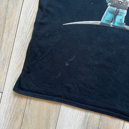 Lego Batman 3: Beyond Gotham 2014 Tee is a black T-shirt made on an Anvil blank. The front features a graphic of Batman in a white and blue suit standing on the moon, holding a Lego flag, with a planet and space station in the background. The back includes the “Batman 3: Beyond Gotham” logo and a Lego logo placed on the upper back area, with small licensing print beneath.