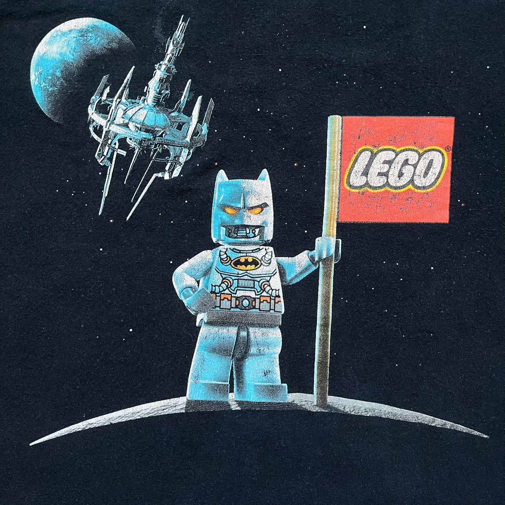 Lego Batman 3: Beyond Gotham 2014 Tee is a black T-shirt made on an Anvil blank. The front features a graphic of Batman in a white and blue suit standing on the moon, holding a Lego flag, with a planet and space station in the background. The back includes the “Batman 3: Beyond Gotham” logo and a Lego logo placed on the upper back area, with small licensing print beneath.