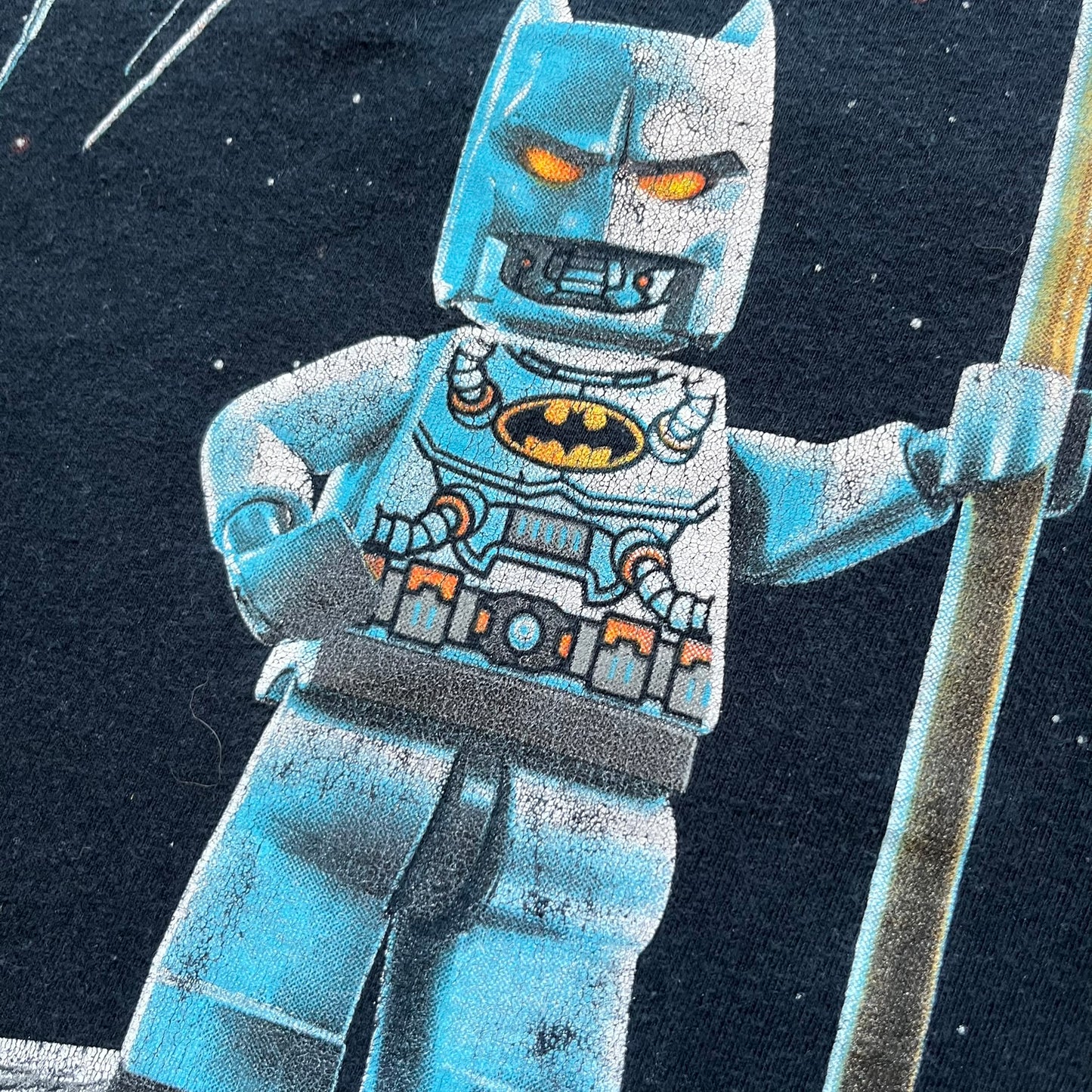 Lego Batman 3: Beyond Gotham 2014 Tee is a black T-shirt made on an Anvil blank. The front features a graphic of Batman in a white and blue suit standing on the moon, holding a Lego flag, with a planet and space station in the background. The back includes the “Batman 3: Beyond Gotham” logo and a Lego logo placed on the upper back area, with small licensing print beneath.