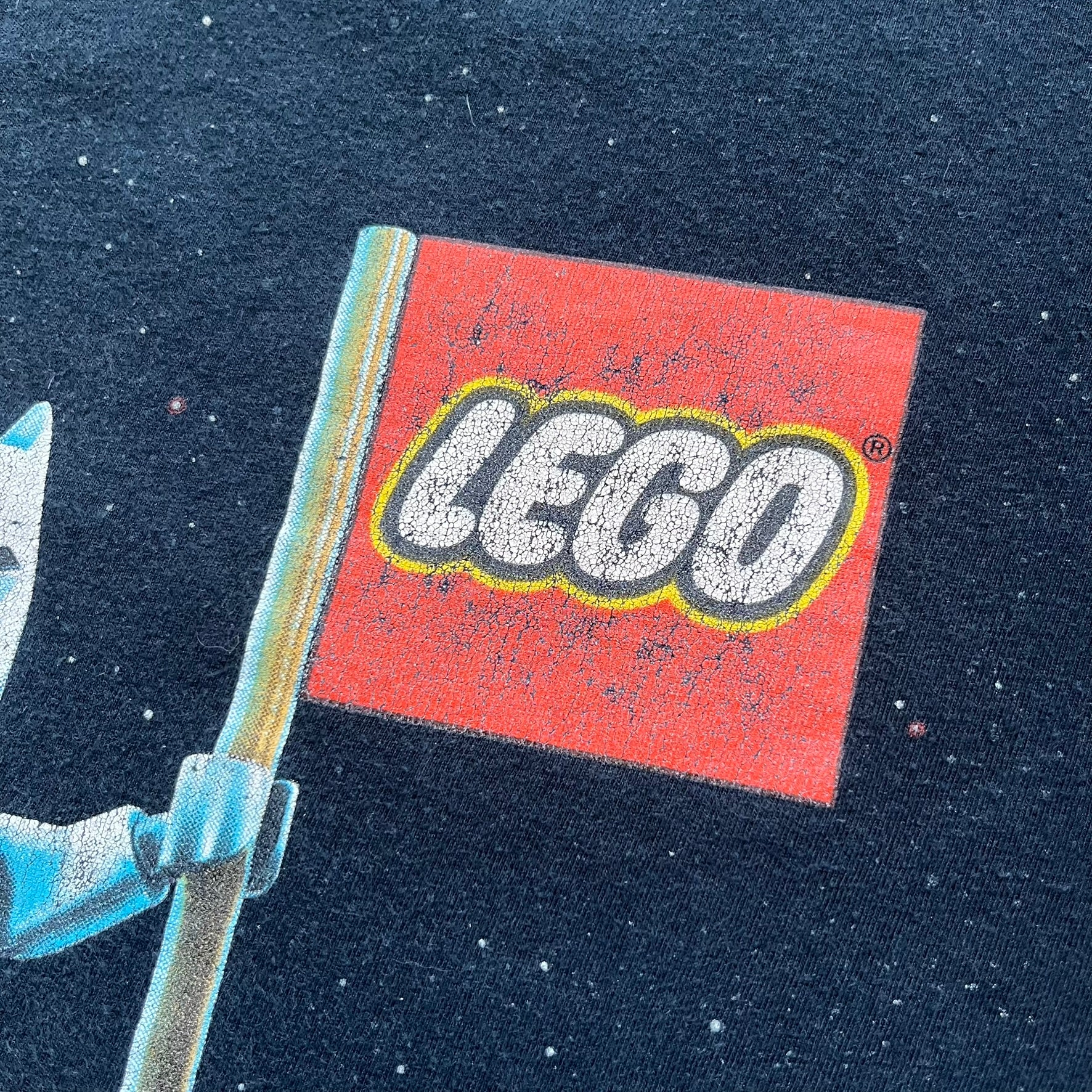 Lego Batman 3: Beyond Gotham 2014 Tee is a black T-shirt made on an Anvil blank. The front features a graphic of Batman in a white and blue suit standing on the moon, holding a Lego flag, with a planet and space station in the background. The back includes the “Batman 3: Beyond Gotham” logo and a Lego logo placed on the upper back area, with small licensing print beneath.