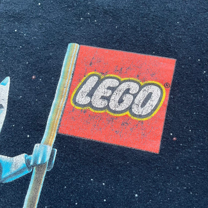 Lego Batman 3: Beyond Gotham 2014 Tee is a black T-shirt made on an Anvil blank. The front features a graphic of Batman in a white and blue suit standing on the moon, holding a Lego flag, with a planet and space station in the background. The back includes the “Batman 3: Beyond Gotham” logo and a Lego logo placed on the upper back area, with small licensing print beneath.