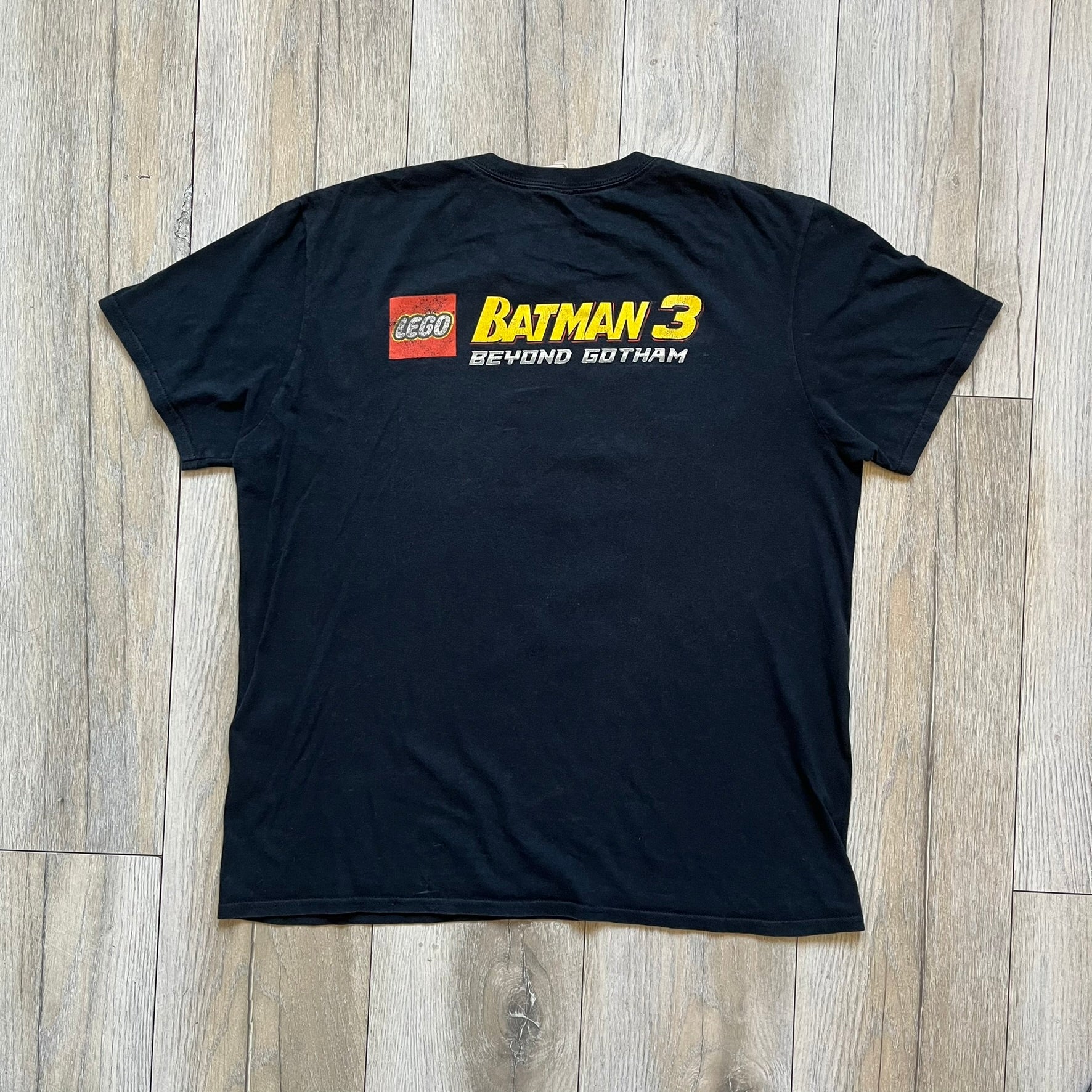 Lego Batman 3: Beyond Gotham 2014 Tee is a black T-shirt made on an Anvil blank. The front features a graphic of Batman in a white and blue suit standing on the moon, holding a Lego flag, with a planet and space station in the background. The back includes the “Batman 3: Beyond Gotham” logo and a Lego logo placed on the upper back area, with small licensing print beneath.