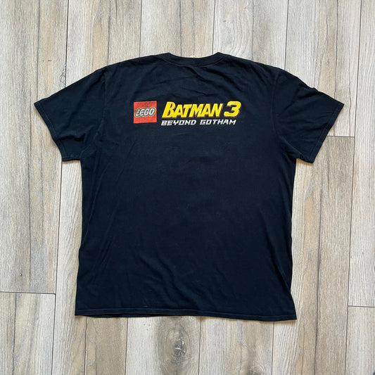 Lego Batman 3: Beyond Gotham 2014 Tee is a black T-shirt made on an Anvil blank. The front features a graphic of Batman in a white and blue suit standing on the moon, holding a Lego flag, with a planet and space station in the background. The back includes the “Batman 3: Beyond Gotham” logo and a Lego logo placed on the upper back area, with small licensing print beneath.