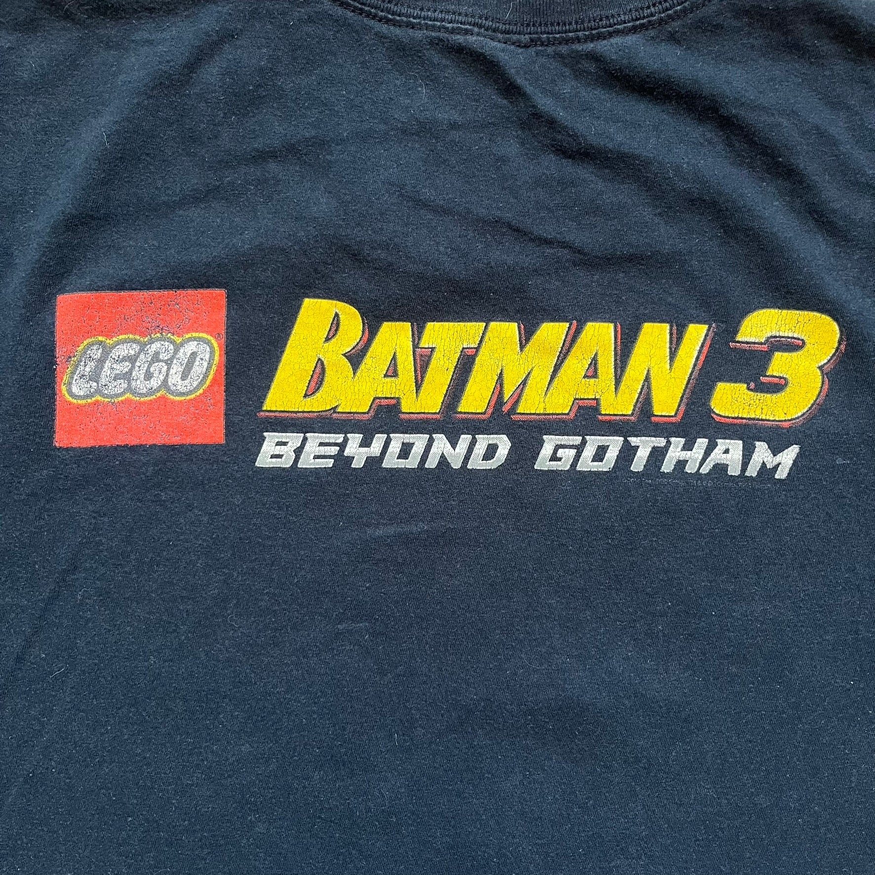 Lego Batman 3: Beyond Gotham 2014 Tee is a black T-shirt made on an Anvil blank. The front features a graphic of Batman in a white and blue suit standing on the moon, holding a Lego flag, with a planet and space station in the background. The back includes the “Batman 3: Beyond Gotham” logo and a Lego logo placed on the upper back area, with small licensing print beneath.