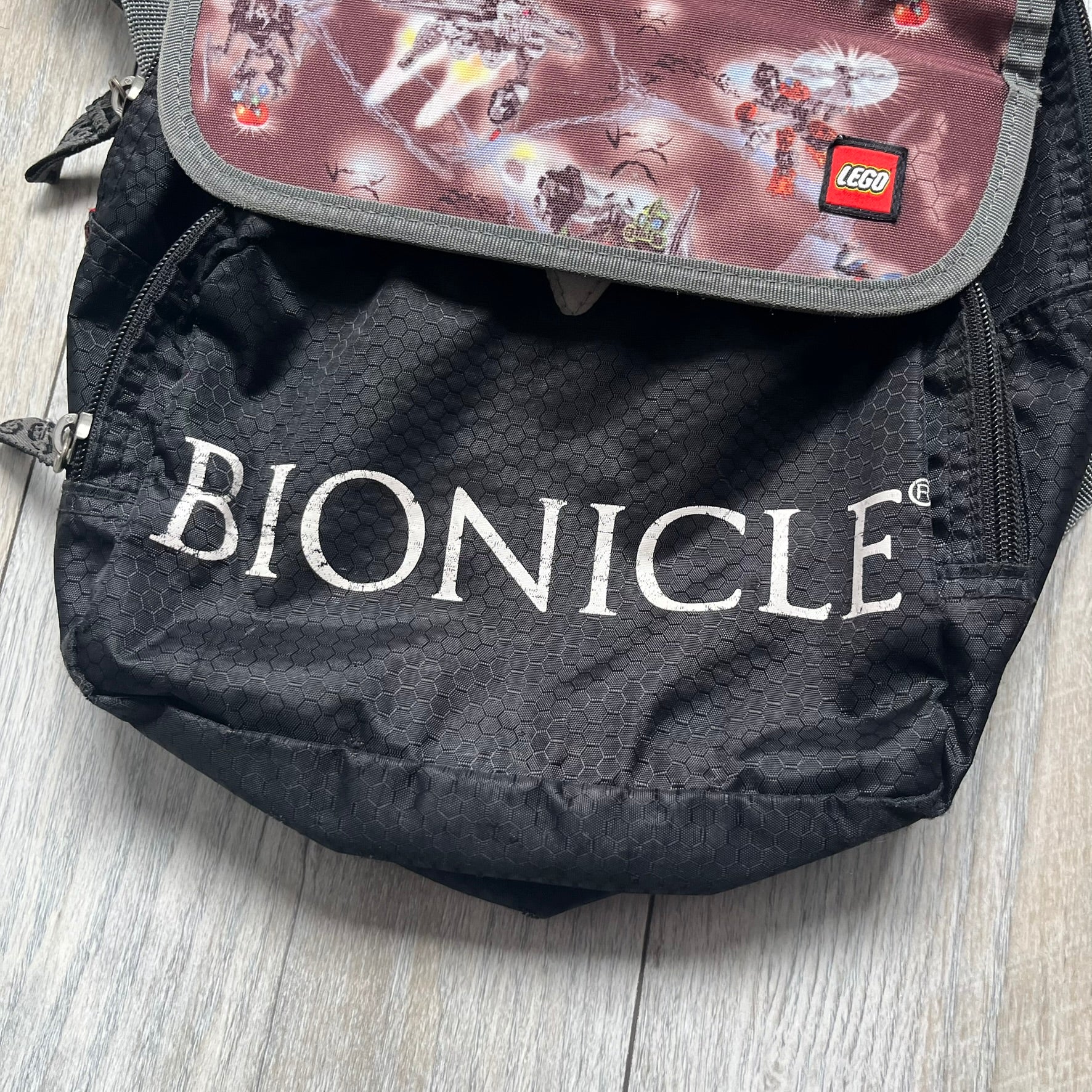 LEGO Bionicle 2009 Small Logo Backpack is a black backpack featuring a large white Bionicle logo on the lower front. The top flap section has a red all-over print with Bionicle characters and a LEGO logo patch. The interior includes a LEGO 2009 licensed tag.