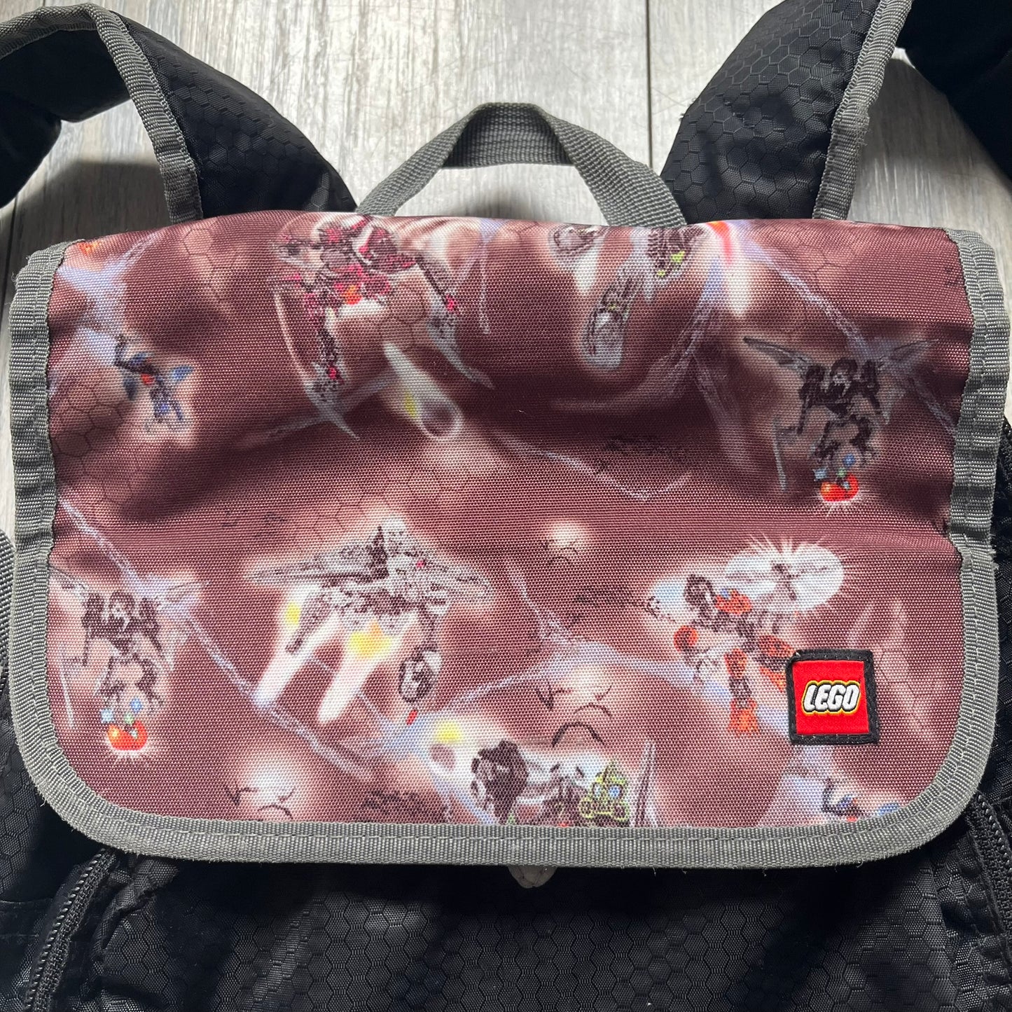 LEGO Bionicle 2009 Small Logo Backpack is a black backpack featuring a large white Bionicle logo on the lower front. The top flap section has a red all-over print with Bionicle characters and a LEGO logo patch. The interior includes a LEGO 2009 licensed tag.