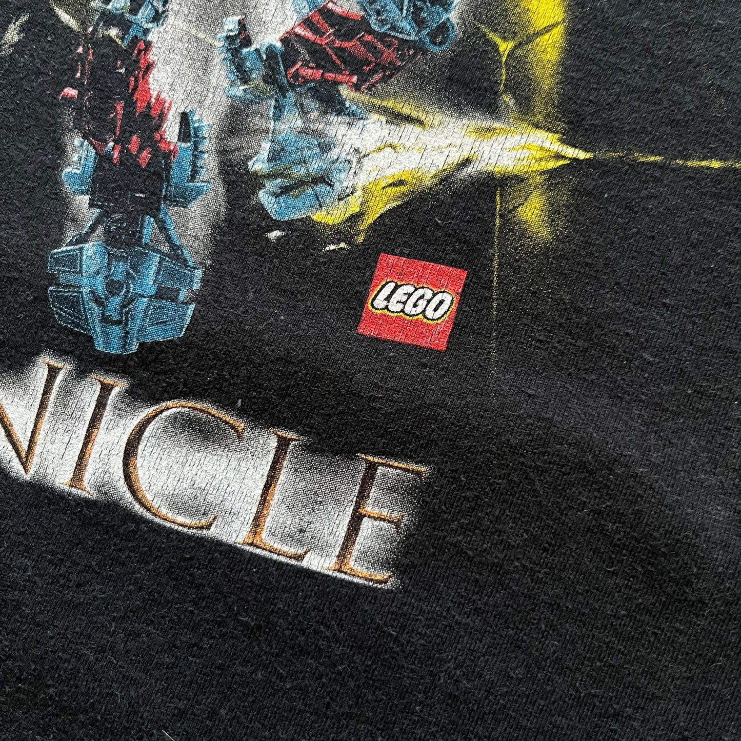 Rare 2000s Lego Bionicle T-shirt showcasing Toa Hordika Vakama from the 2005 set. Features the glowing Bionicle logo under the Binicle, small Lego branding and a naturally sunfaded black wash. A collectible tee that perfectly captures early-2000s nostalgia.