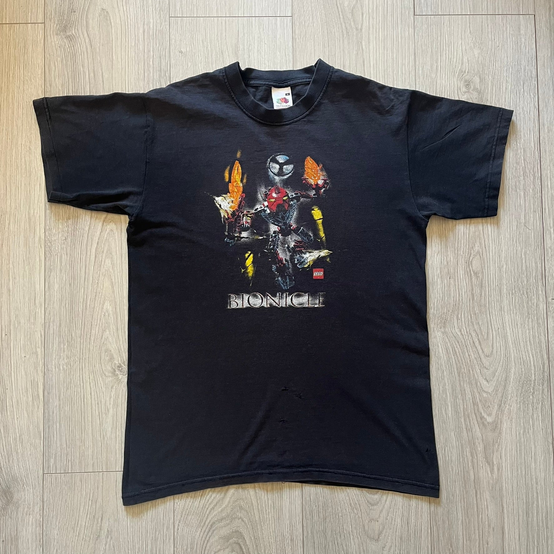 Rare 2000s Lego Bionicle T-shirt showcasing Toa Hordika Vakama from the 2005 set. Features the glowing Bionicle logo under the Binicle, small Lego branding and a naturally sunfaded black wash. A collectible tee that perfectly captures early-2000s nostalgia.