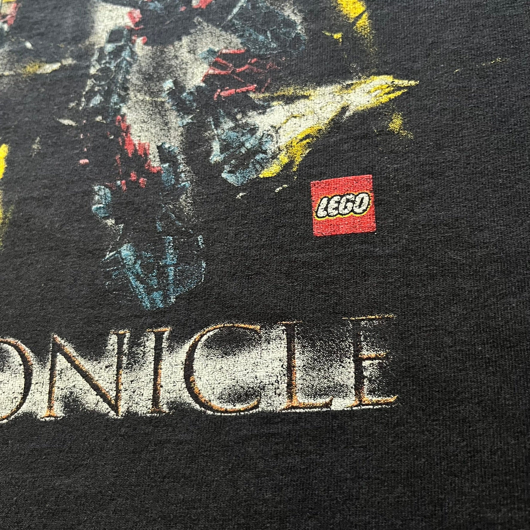 Rare 2000s Lego Bionicle T-shirt showcasing Toa Hordika Vakama from the 2005 set. Features the glowing Bionicle logo under the Binicle, small Lego branding and a naturally sunfaded black wash. A collectible tee that perfectly captures early-2000s nostalgia.