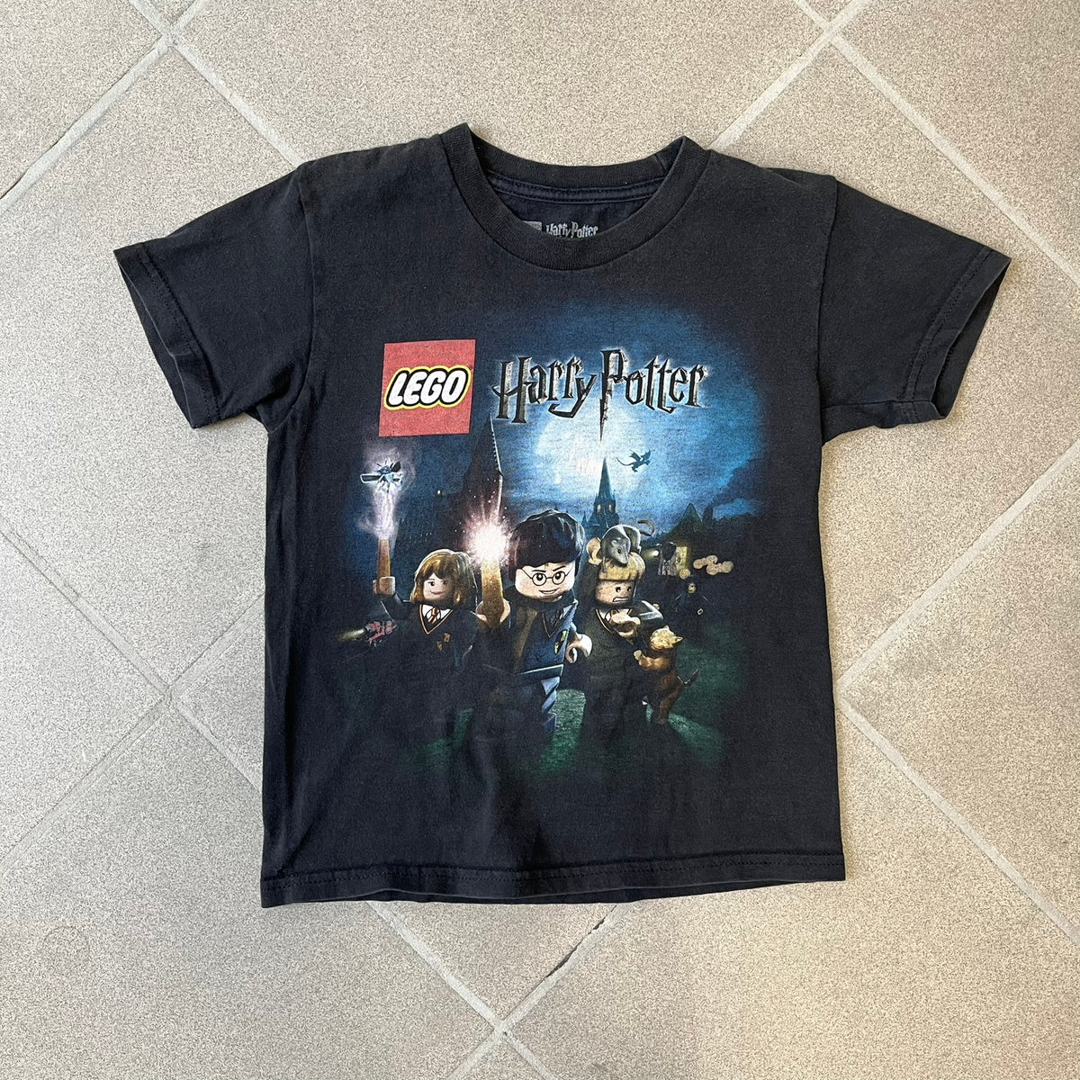 Lego Harry Potter: Years 1-4 Game 2011 Cover Tee is a black kids' T-shirt made from 100% cotton, featuring the game's cover artwork with classic characters and the Lego Harry Potter logo on the front. It carries a licensed 2011 Warner Bros. neck tag.