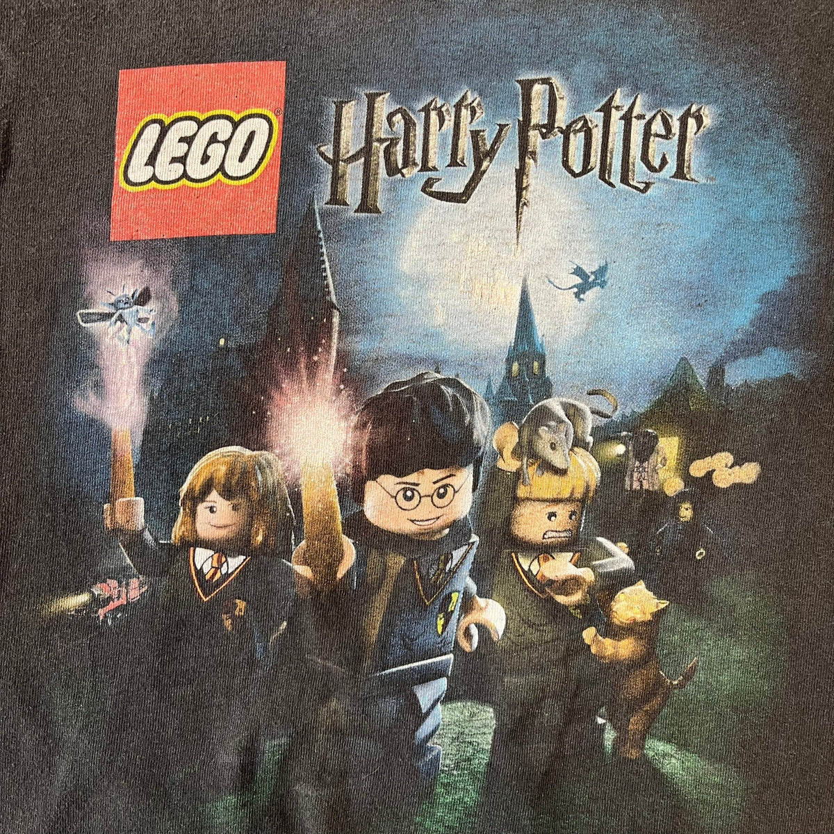 Lego Harry Potter: Years 1-4 Game 2011 Cover Tee is a black kids' T-shirt made from 100% cotton, featuring the game's cover artwork with classic characters and the Lego Harry Potter logo on the front. It carries a licensed 2011 Warner Bros. neck tag.