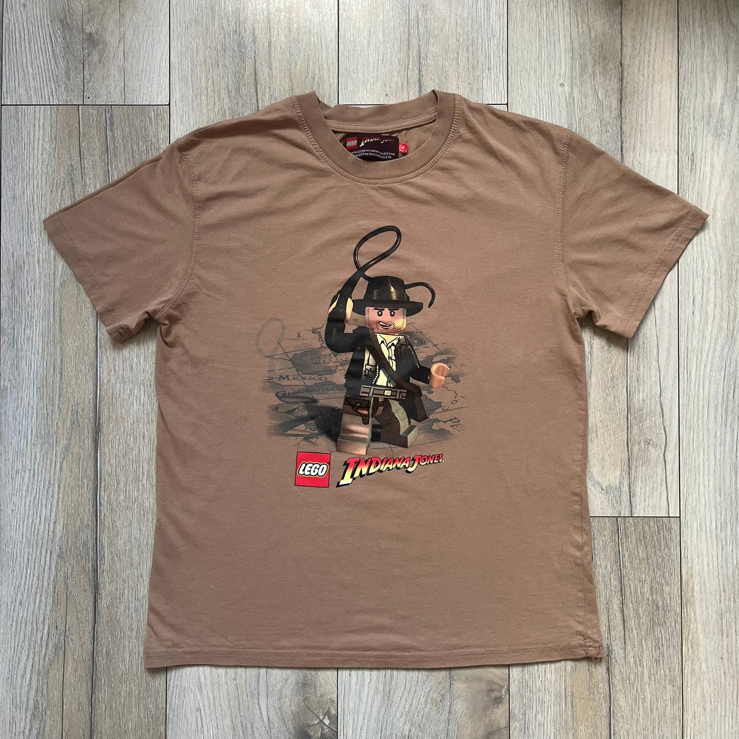 Lego Indiana Jones 2009 Brown Tee is a light brown short-sleeve T-shirt featuring a front graphic of Lego Indiana Jones swinging a whip with the Lego Indiana Jones logo beneath. A faint map appears behind the character, and the neck tag displays 2009 licensing. The graphic is sourced from the original Lego Indiana Jones game.