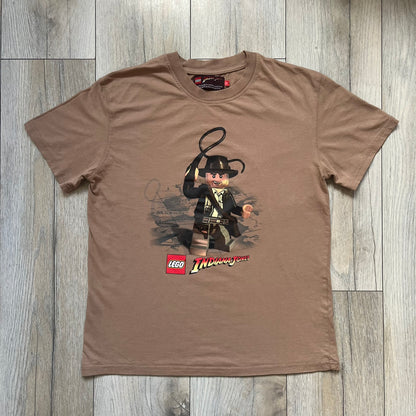 Lego Indiana Jones 2009 Brown Tee is a light brown short-sleeve T-shirt featuring a front graphic of Lego Indiana Jones swinging a whip with the Lego Indiana Jones logo beneath. A faint map appears behind the character, and the neck tag displays 2009 licensing. The graphic is sourced from the original Lego Indiana Jones game.