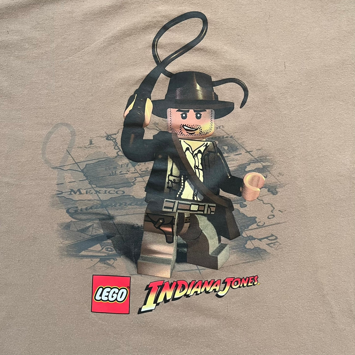 Lego Indiana Jones 2009 Brown Tee is a light brown short-sleeve T-shirt featuring a front graphic of Lego Indiana Jones swinging a whip with the Lego Indiana Jones logo beneath. A faint map appears behind the character, and the neck tag displays 2009 licensing. The graphic is sourced from the original Lego Indiana Jones game.