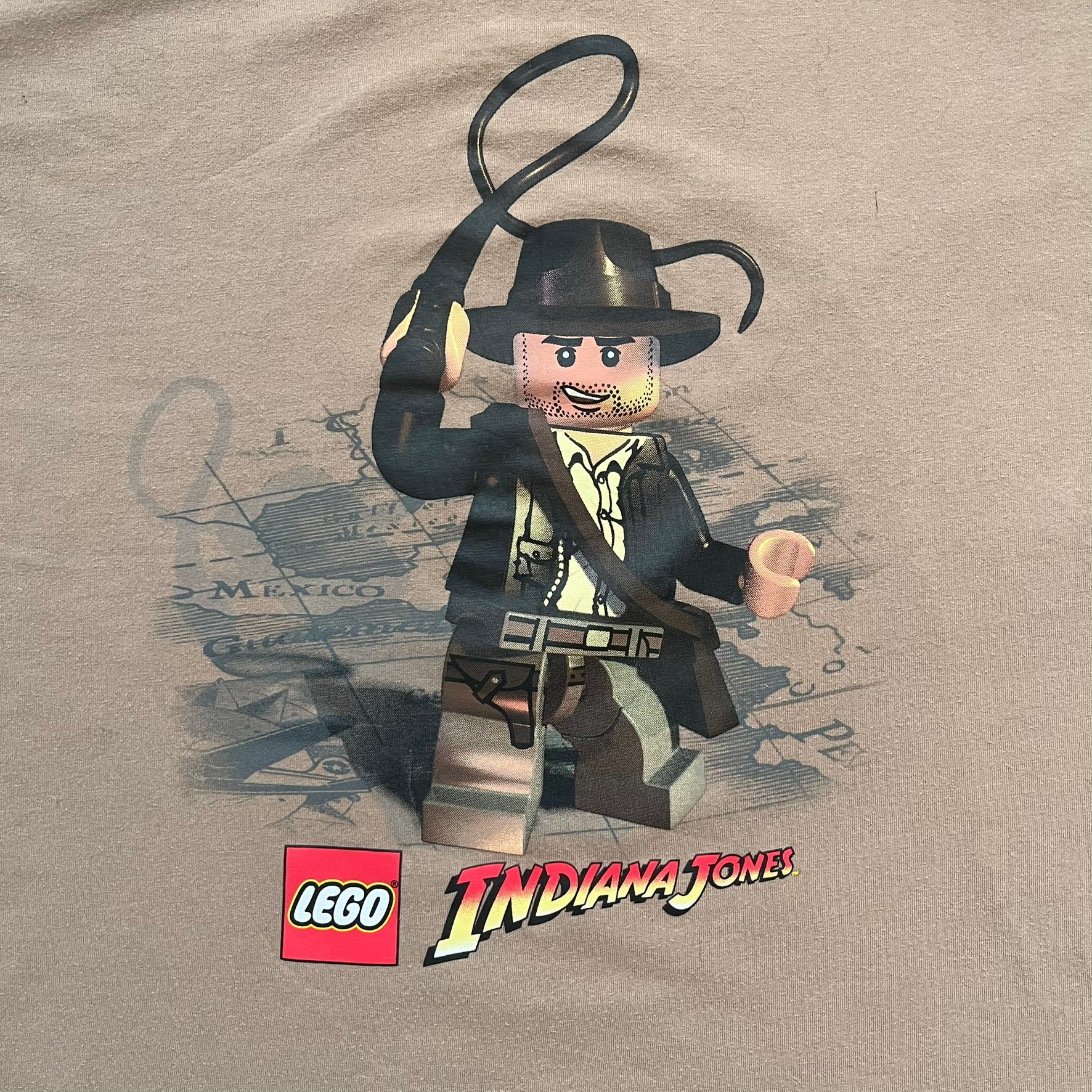 Lego Indiana Jones 2009 Brown Tee is a light brown short-sleeve T-shirt featuring a front graphic of Lego Indiana Jones swinging a whip with the Lego Indiana Jones logo beneath. A faint map appears behind the character, and the neck tag displays 2009 licensing. The graphic is sourced from the original Lego Indiana Jones game.