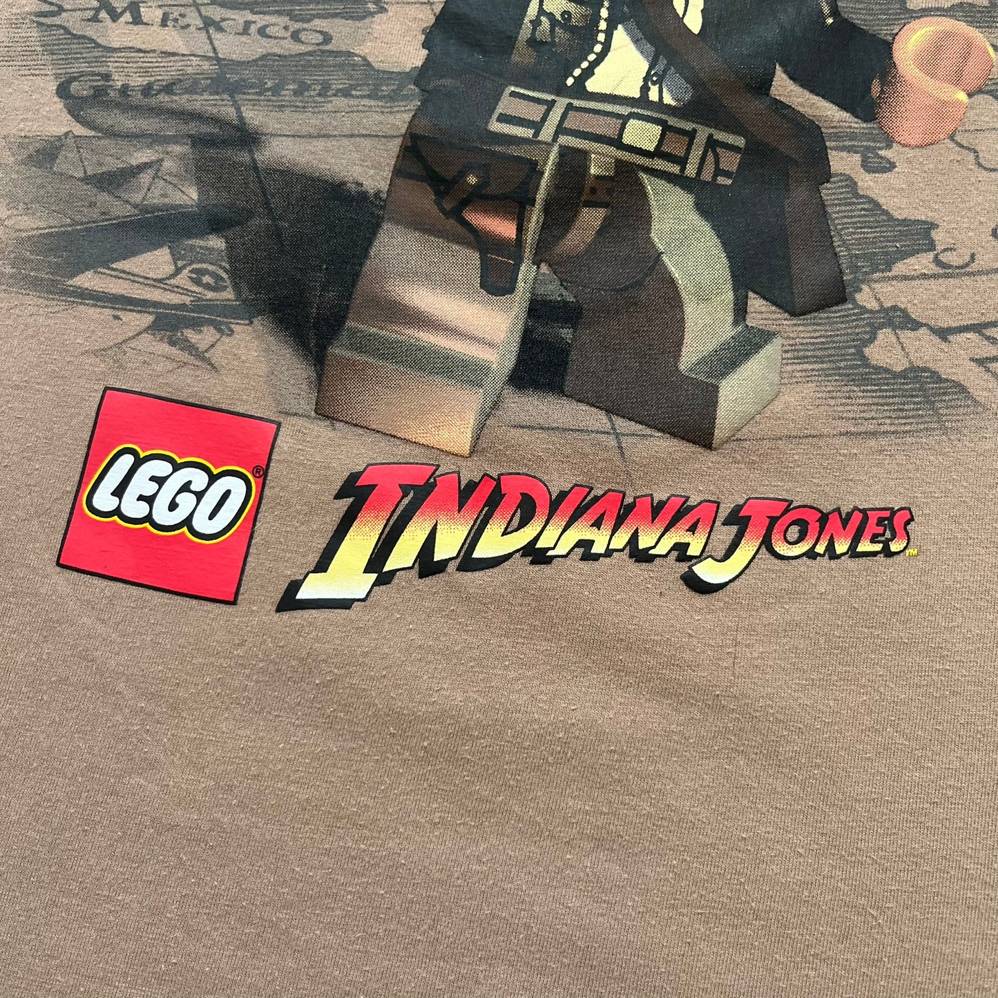 Lego Indiana Jones 2009 Brown Tee is a light brown short-sleeve T-shirt featuring a front graphic of Lego Indiana Jones swinging a whip with the Lego Indiana Jones logo beneath. A faint map appears behind the character, and the neck tag displays 2009 licensing. The graphic is sourced from the original Lego Indiana Jones game.