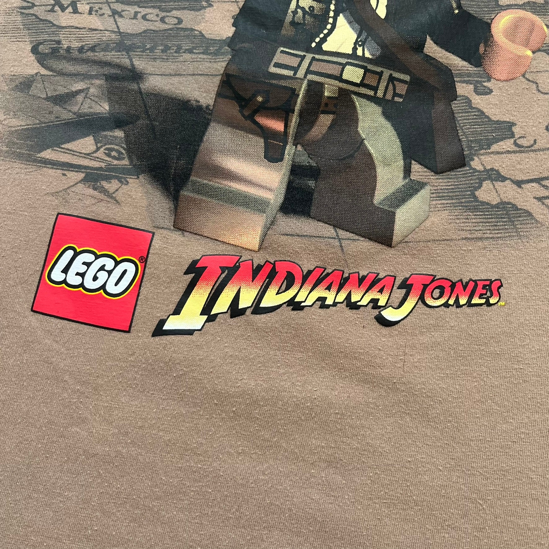 Lego Indiana Jones 2009 Brown Tee is a light brown short-sleeve T-shirt featuring a front graphic of Lego Indiana Jones swinging a whip with the Lego Indiana Jones logo beneath. A faint map appears behind the character, and the neck tag displays 2009 licensing. The graphic is sourced from the original Lego Indiana Jones game.