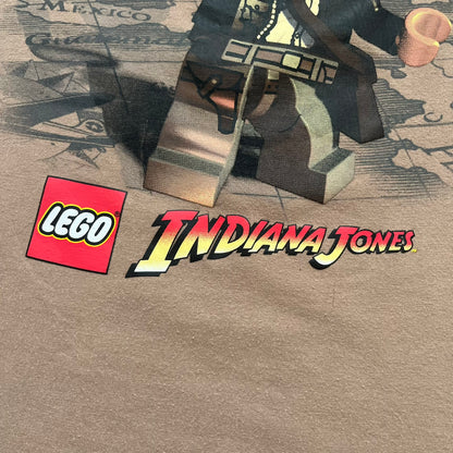 Lego Indiana Jones 2009 Brown Tee is a light brown short-sleeve T-shirt featuring a front graphic of Lego Indiana Jones swinging a whip with the Lego Indiana Jones logo beneath. A faint map appears behind the character, and the neck tag displays 2009 licensing. The graphic is sourced from the original Lego Indiana Jones game.