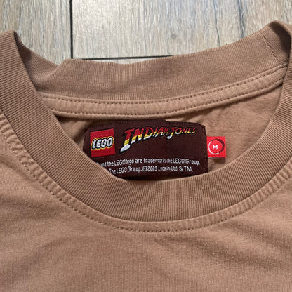 Lego Indiana Jones 2009 Brown Tee is a light brown short-sleeve T-shirt featuring a front graphic of Lego Indiana Jones swinging a whip with the Lego Indiana Jones logo beneath. A faint map appears behind the character, and the neck tag displays 2009 licensing. The graphic is sourced from the original Lego Indiana Jones game.
