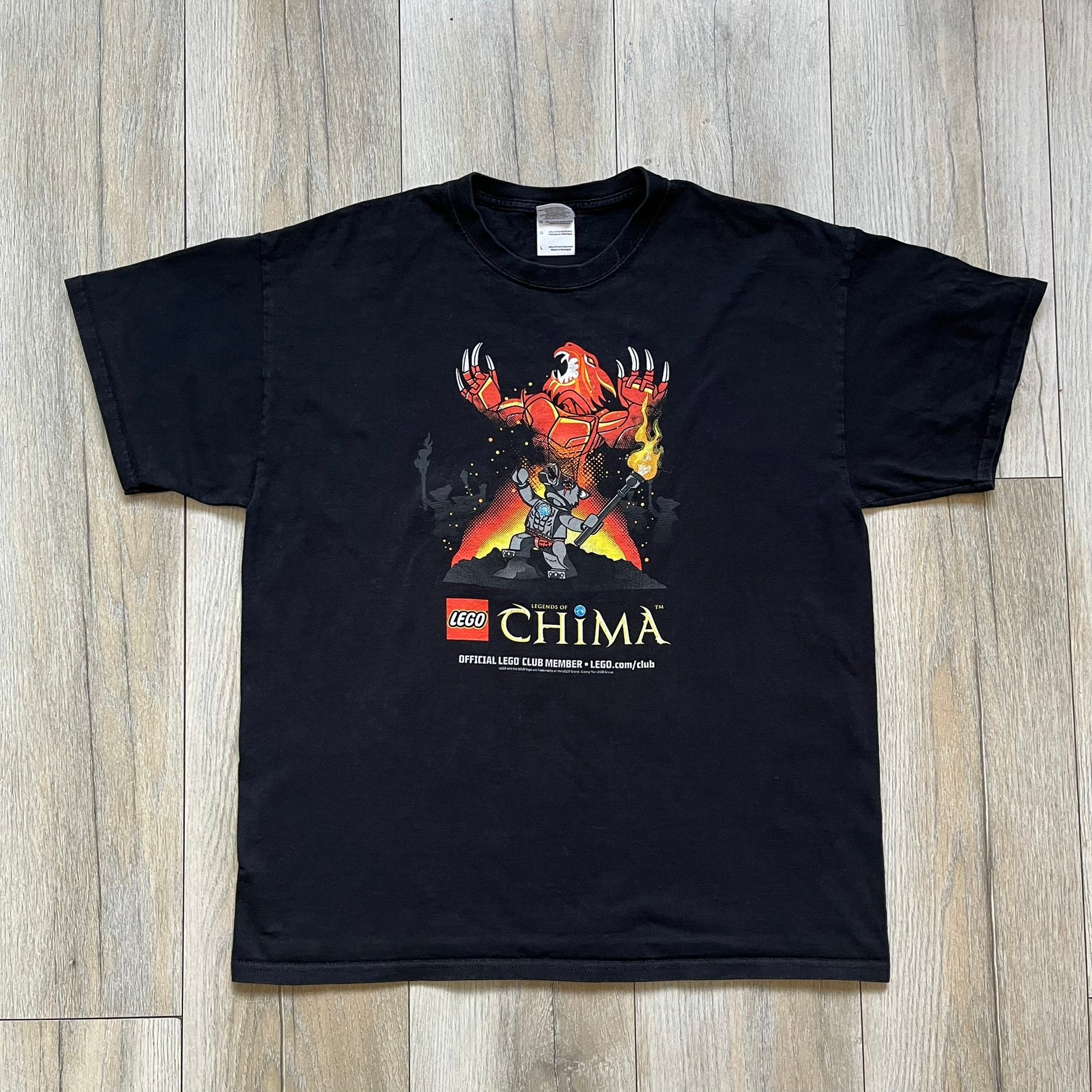 Lego Legends of Chima 2014 Black Tee is a black T-shirt made on a Gildan Ultra Cotton blank. The front features Winzar holding a torch with a large red character behind him and additional faded grey characters in the background. Below the graphic are the Lego Legends of Chima logo, “Official Lego Club Member” text, website link, and small 2014 licensing print.