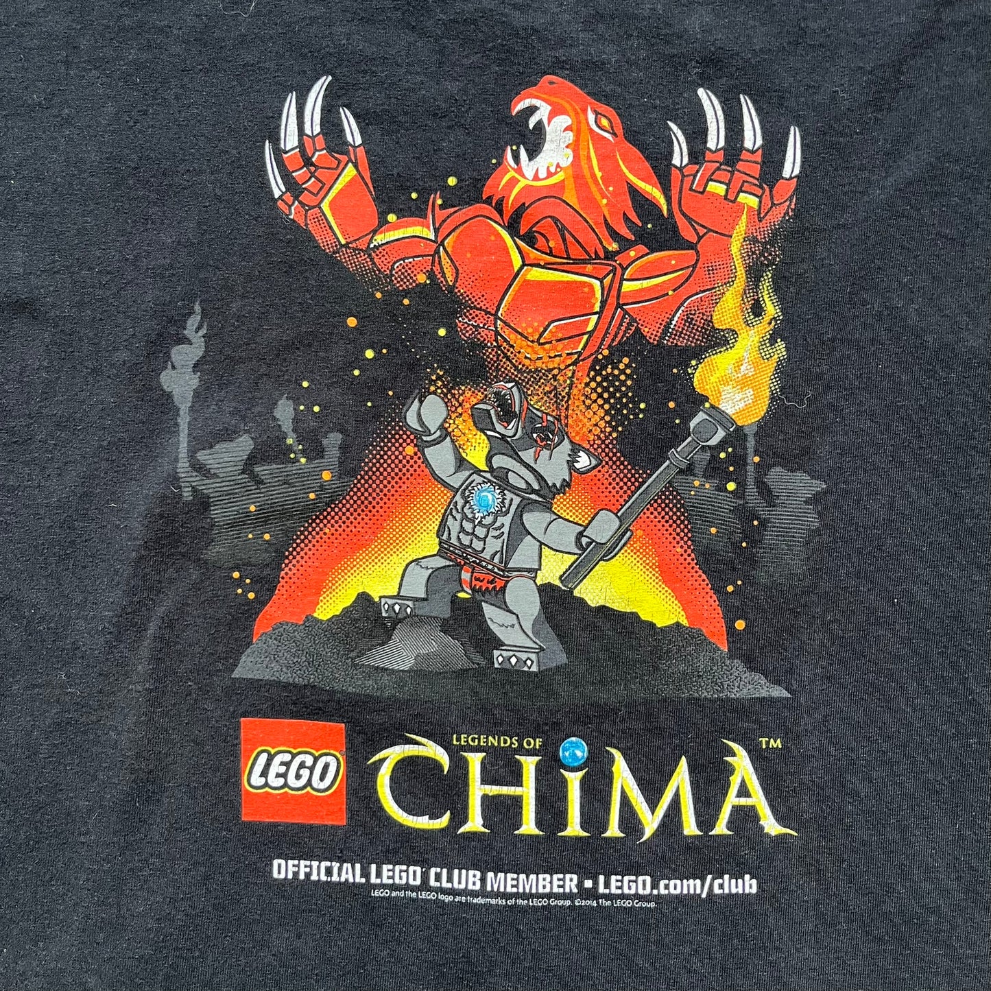 Lego Legends of Chima 2014 Black Tee is a black T-shirt made on a Gildan Ultra Cotton blank. The front features Winzar holding a torch with a large red character behind him and additional faded grey characters in the background. Below the graphic are the Lego Legends of Chima logo, “Official Lego Club Member” text, website link, and small 2014 licensing print.