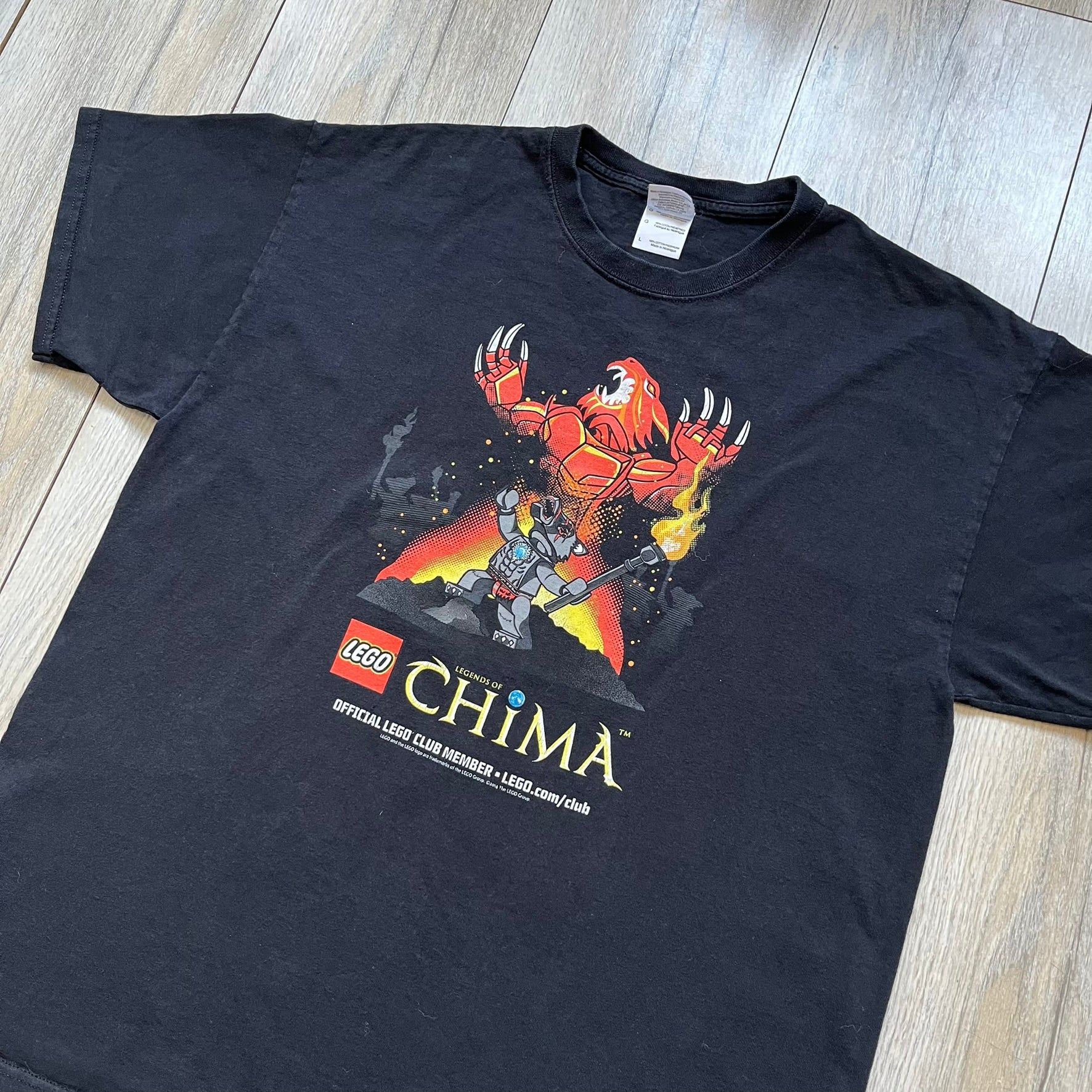 Lego Legends of Chima 2014 Black Tee is a black T-shirt made on a Gildan Ultra Cotton blank. The front features Winzar holding a torch with a large red character behind him and additional faded grey characters in the background. Below the graphic are the Lego Legends of Chima logo, “Official Lego Club Member” text, website link, and small 2014 licensing print.