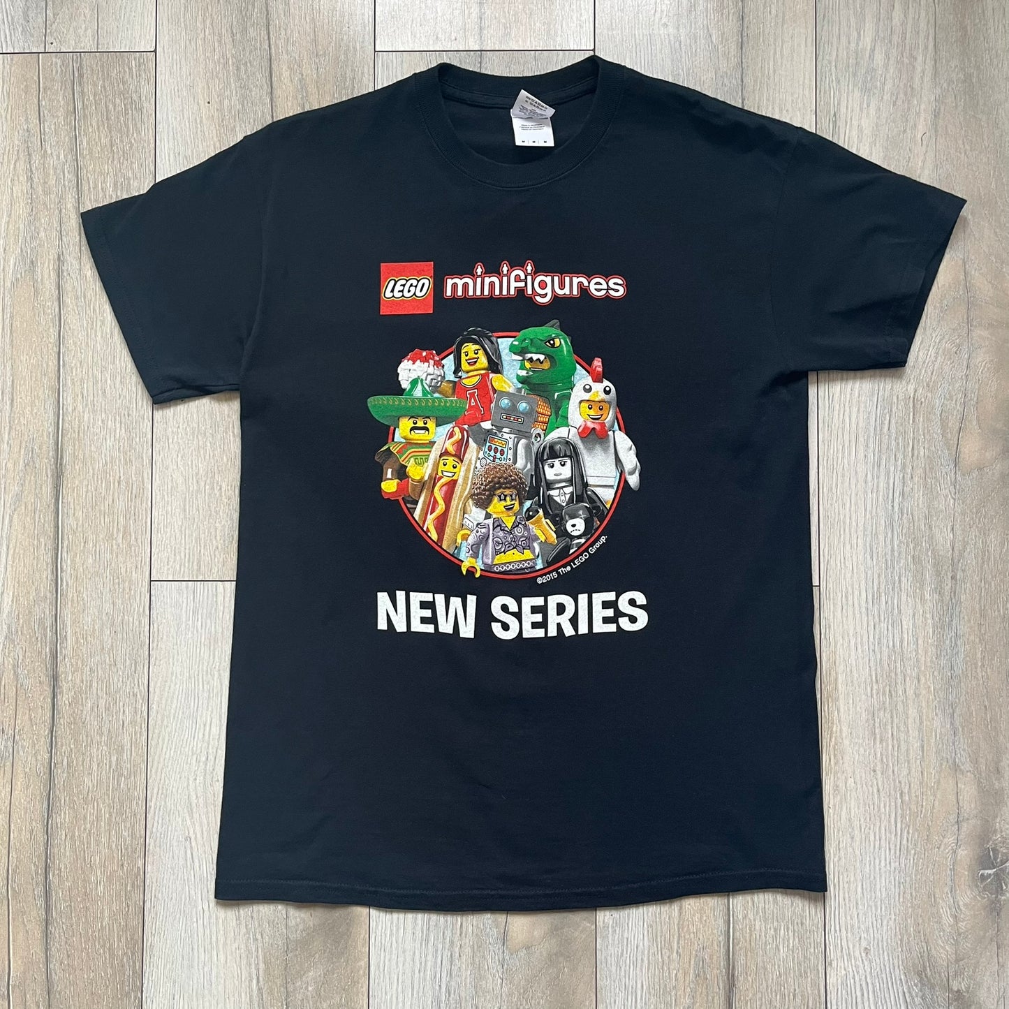 LEGO Minifigures “New Series” 2015 Tee is a black T-shirt featuring front and back prints. The front displays “LEGO Minifigures” text above eight characters arranged in a circle, with 2015 LEGO licensing and “New Series” text below. The back repeats the logo with a yellow minifigure in the center and “ASK ME” text underneath, likely produced as an employee promotional item.